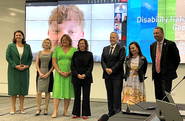 Disability Reform Ministers stand in front of a large screen hosting other council meeting attendees