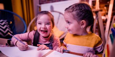 Two children at a table. One child is smiling while writing on a piece of paper